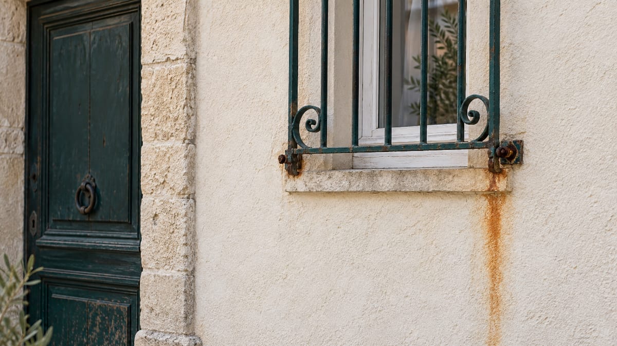 Coulure de rouille sur une façade enduite blanche sous une grille de fenêtre en fer